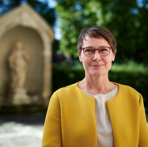 Birte Graue, Frau mit Kurzhaarfrisur und Brille in senffarbenem Oberteil lächelt in die Kamera. Im Hintergrund verschwommen eine Sandsteinstatue.