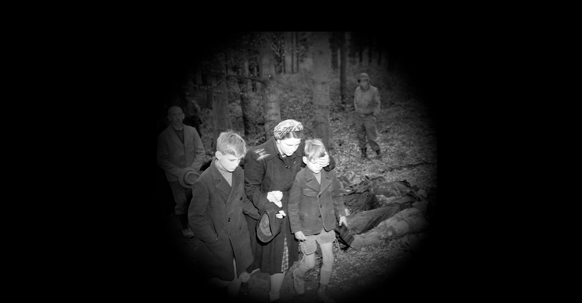 Eine junge Frau führt zwei Kinder an einer Reihe von Leichen vorbei, die auf dem Waldboden liegen, im Hintergrund steht ein amerikanischer Soldat.
