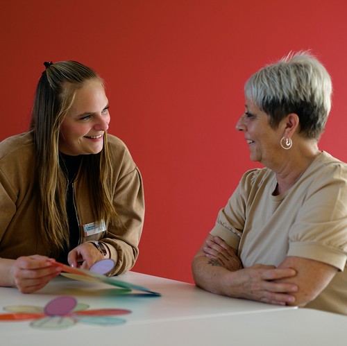 Junge Frau unterhält sich mit älterer Frau, zeigt auf ein buntes Arbeitsblatt in Form einer Blume